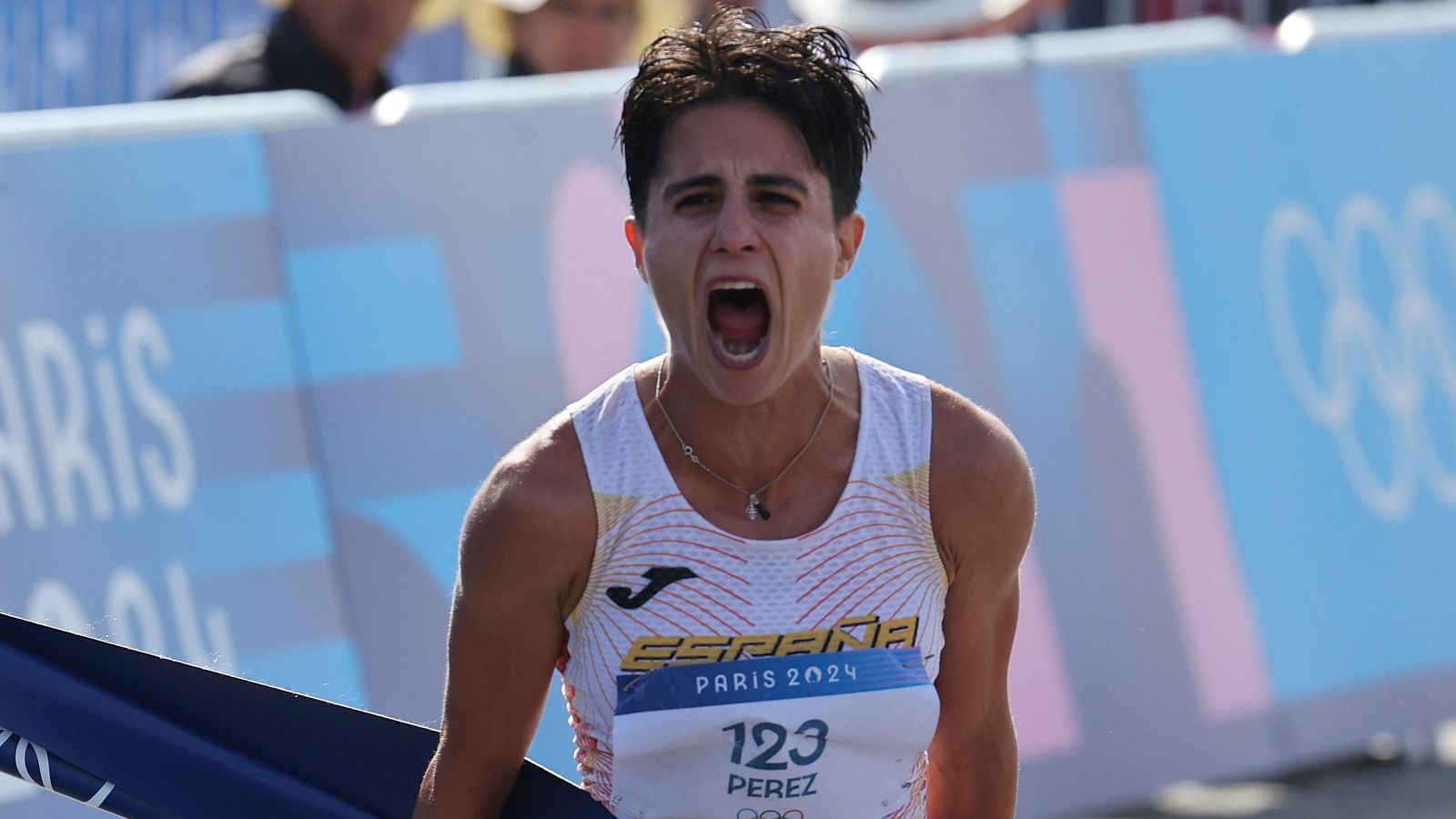 Atleta cruzando la meta con expresión de triunfo; camiseta blanca con detalles rojos y dorados, dorsal 123; fondo con valla publicitaria de París 2024.