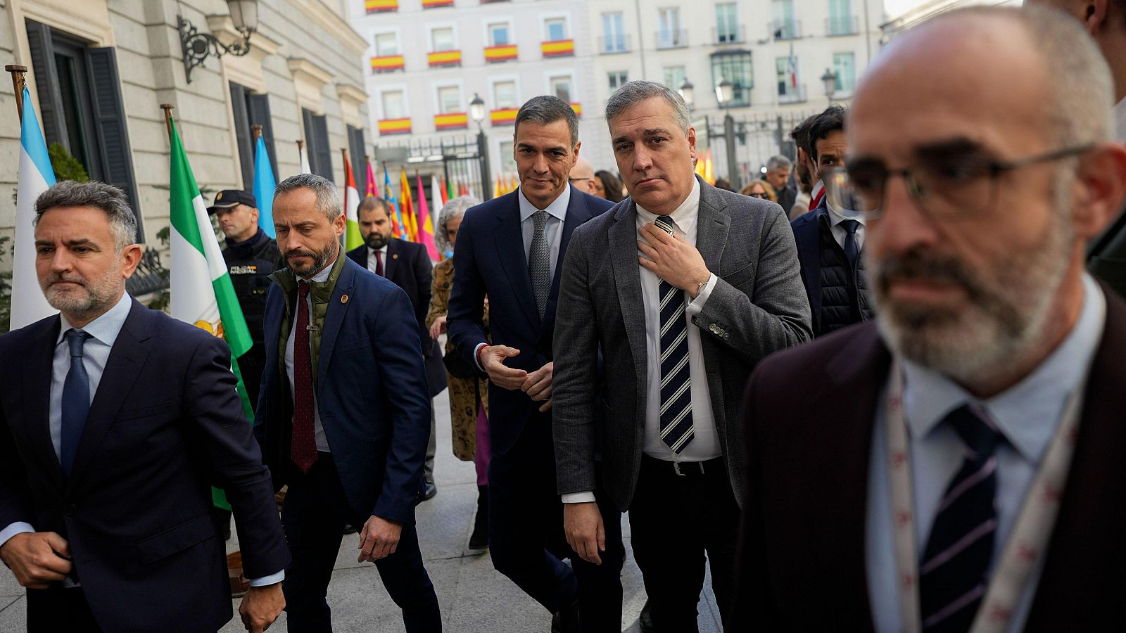 Grupo de personas en traje, posiblemente políticos, en exterior. Un hombre con barba y gafas en primer plano; otros, ligeramente desenfocados, al fondo.  Ambiente formal.