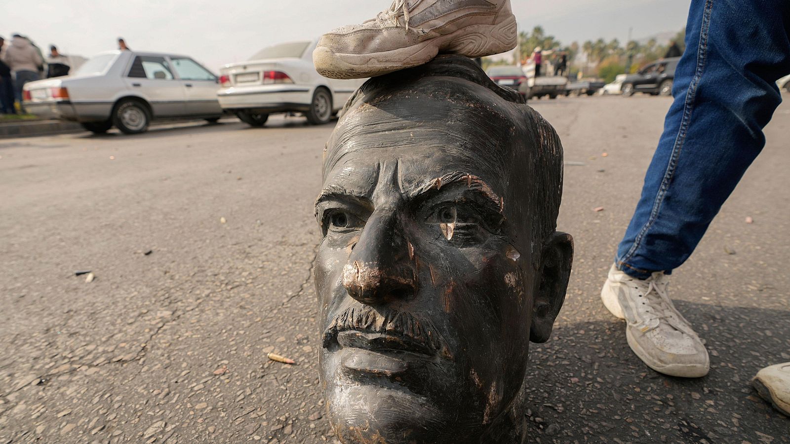 Busto roto en el suelo, pisado por una zapatilla blanca.  Fondo con coches aparcados.  Posible acto de desafío.