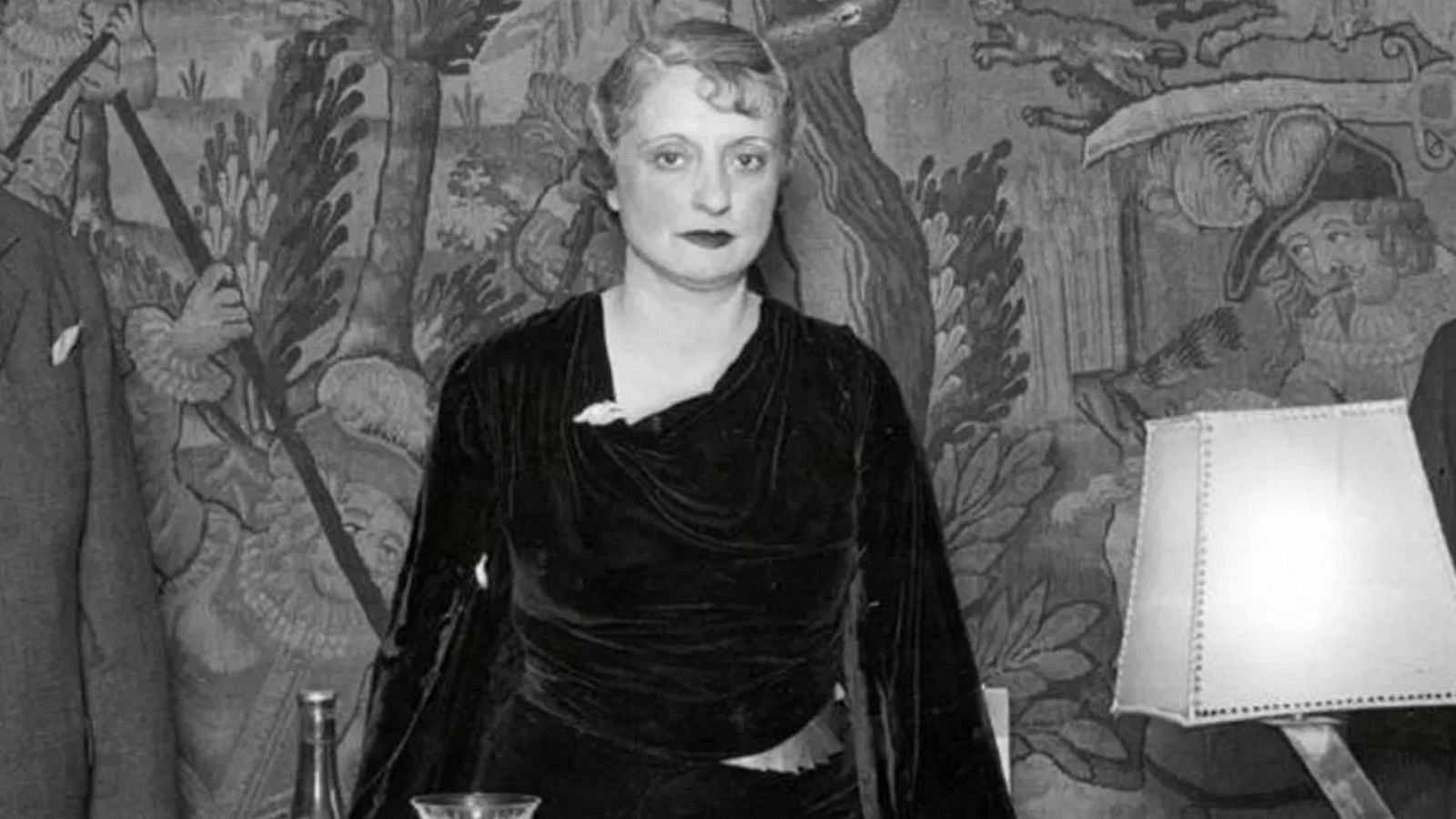 Retrato en blanco y negro de Mari Luz Morales con vestido oscuro, sentada en un evento social con una copa y botella en la mesa cercana. Fondo con tapiz y otras personas.