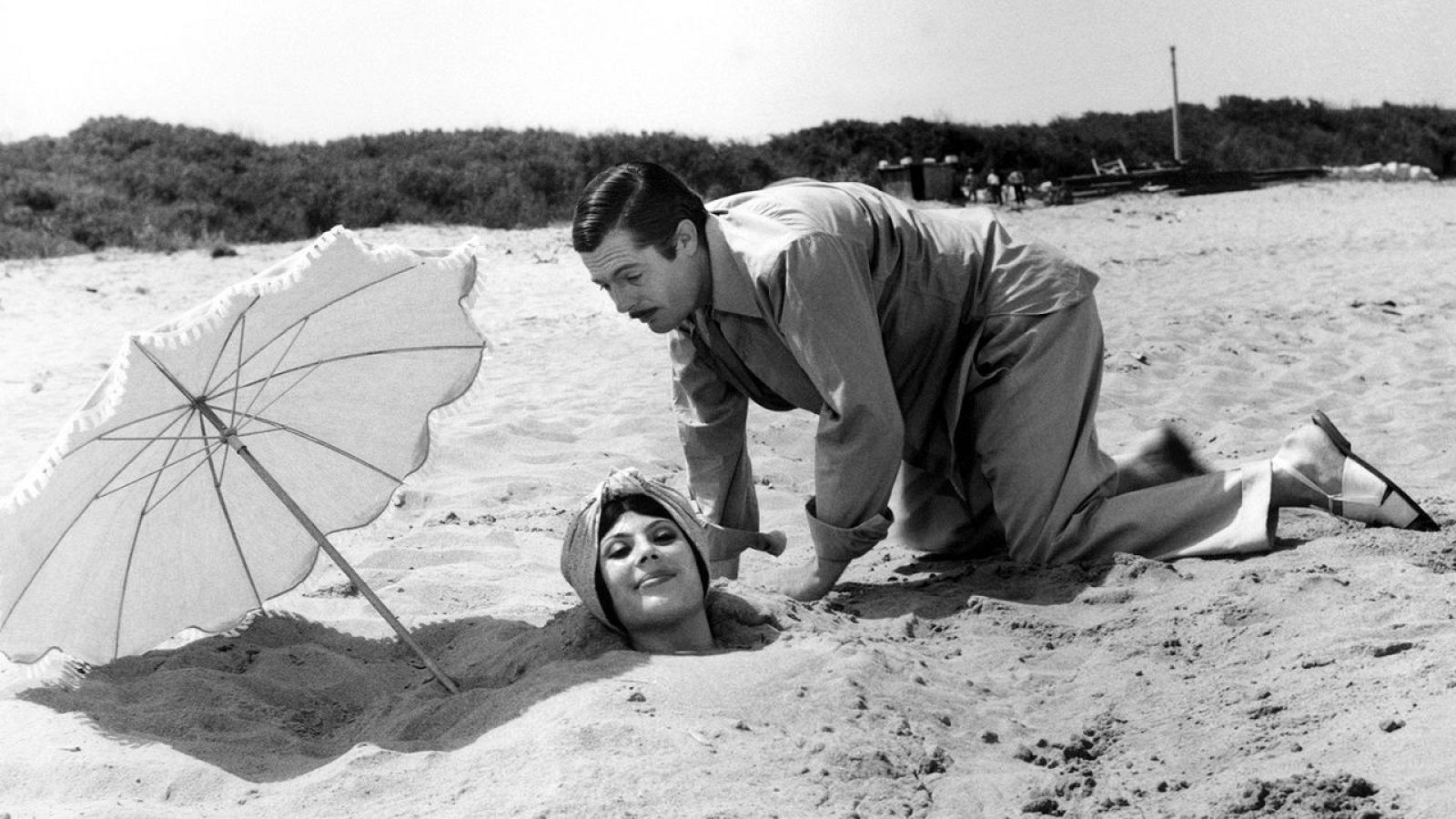Escena de playa en blanco y negro: Marcello Mastroianni arrodillado junto a Daniela Rocca, cuya cabeza asoma de la arena; una sombrilla blanca al fondo.