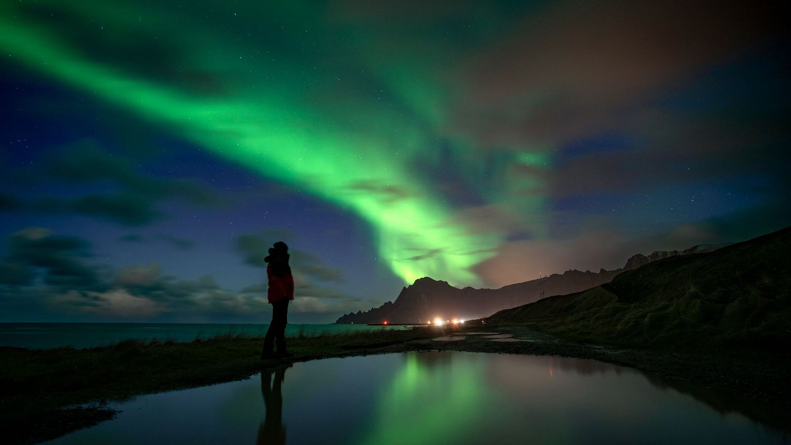Aurora boreal sobre Noruega: luces verdes ondulantes en el cielo nocturno, reflejadas en un charco de agua.  Una persona observa el fenómeno desde la orilla, con montañas al fondo.