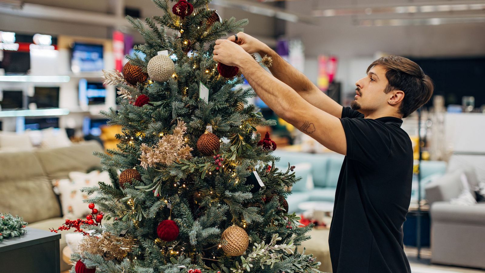Joven colocando adornos navideños (rojos y dorados) en un árbol de Navidad, en un espacio que parece una tienda o almacén.