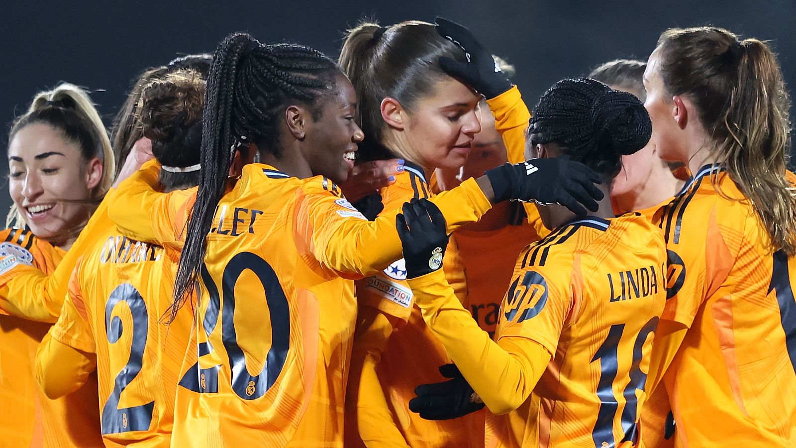 Celebración de jugadoras del Real Madrid tras un gol o victoria en partido de la UEFA Women's Champions League. Se ven dorsales 2, 18 (LINDA) y 20 (OLLET) en camisetas naranjas.