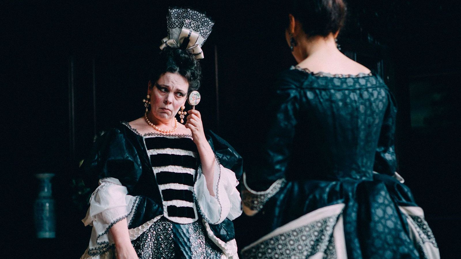 Retrato de Olivia Colman con un vestido negro de época,  peinado recogido y un espejo en la mano; expresión seria.