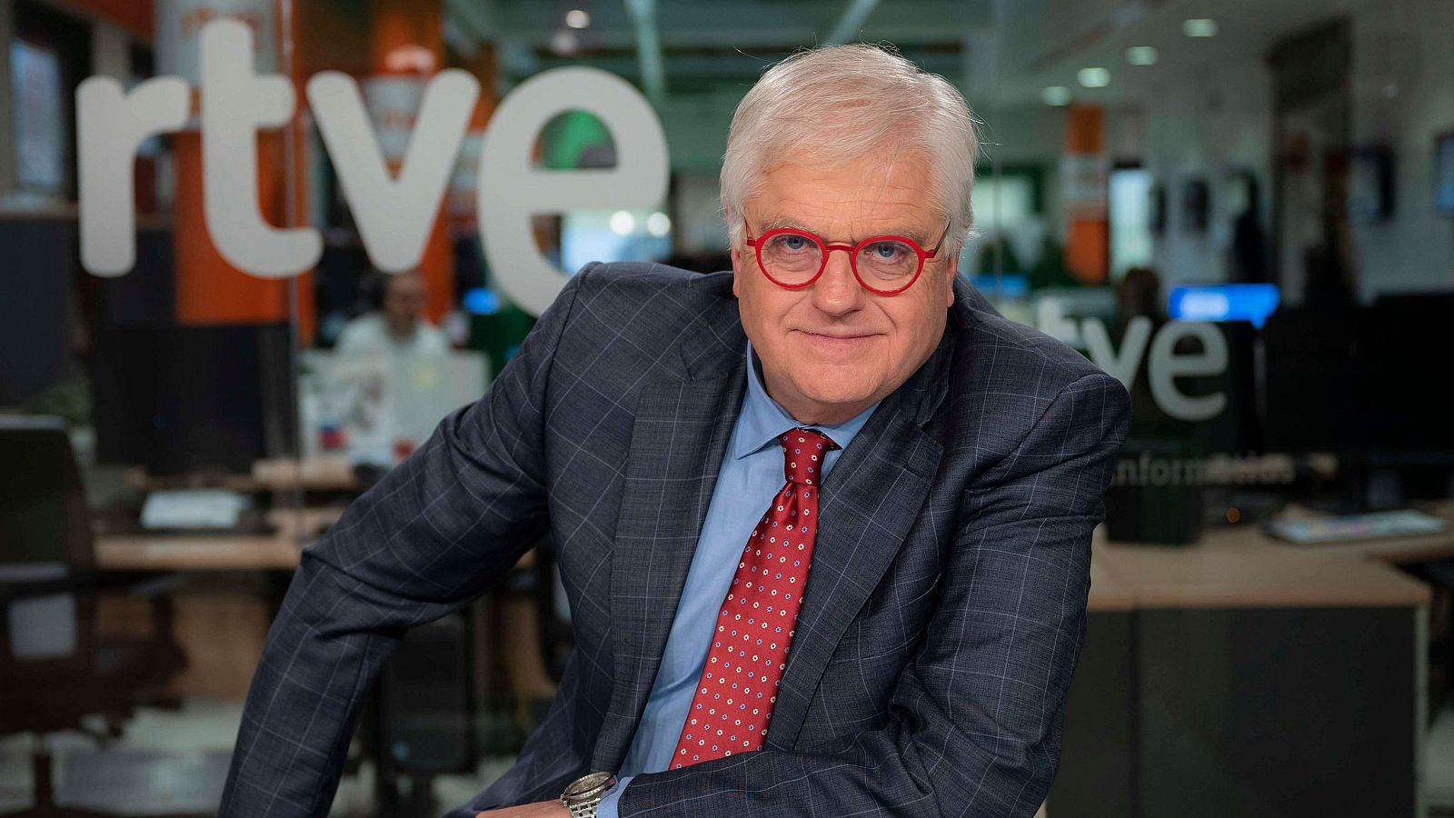 Lluís Falgàs, presentador d''Aquí parlem' de TVE Catalunya, apareix en una imatge promocional. Vesteix una jaqueta grisa a quadres, camisa blava i corbata granat amb lunars blancs. El fons mostra un estudi de televisió.