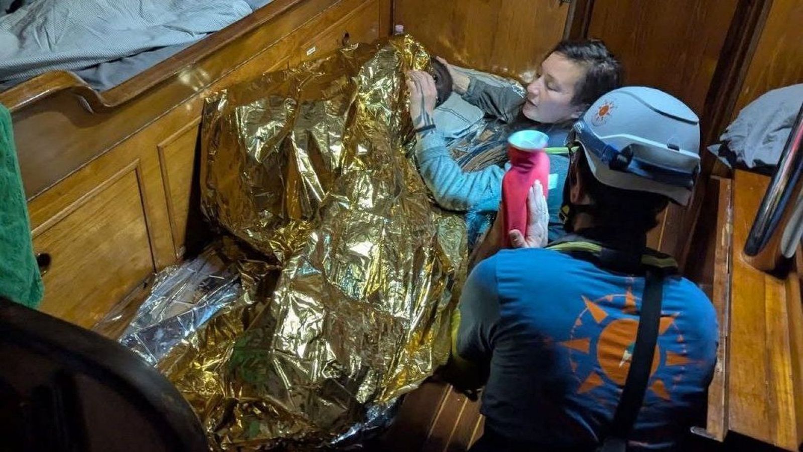 Niña envuelta en manta térmica dorada recibe atención médica; una persona le acerca una bolsa de agua caliente, otra lleva casco de rescate.