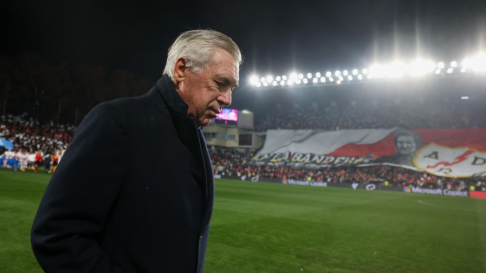 Imagen de Carlo Ancelotti al borde del campo, con la cabeza baja.  Aficionados en la grada y la iluminación del estadio son visibles.  Contexto: tensión entre optimismo y expectativas.