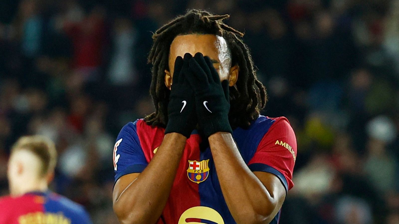 Futbolista con camiseta azulgrana y guantes negros se cubre el rostro mostrando decepción.  Otro jugador se ve al fondo.