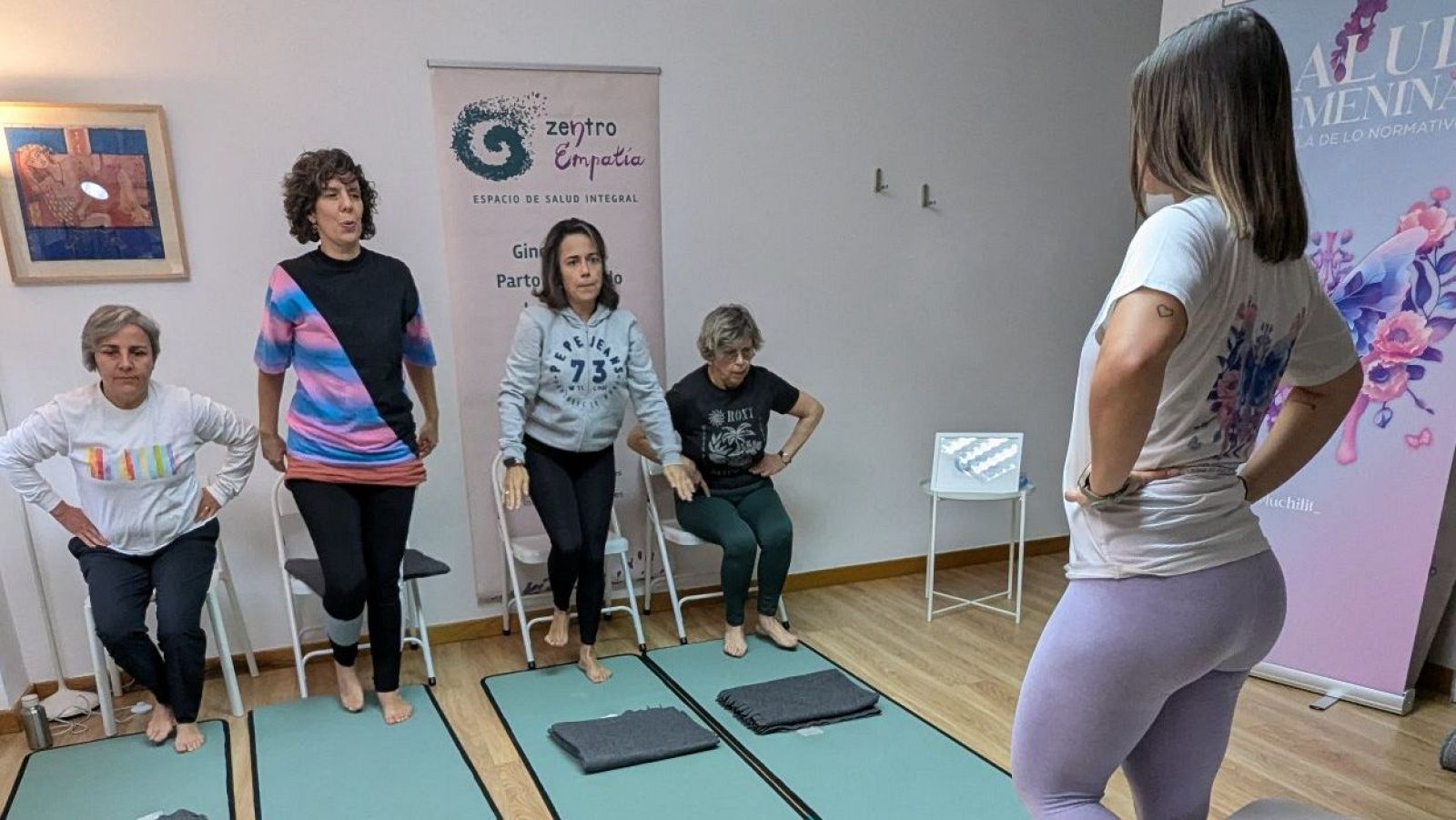 Mujeres realizan ejercicios de fuerza en un taller de climaterio.  Se observa una instructora y participantes con ropa deportiva, en una sala con colchonetas verdes y carteles informativos.