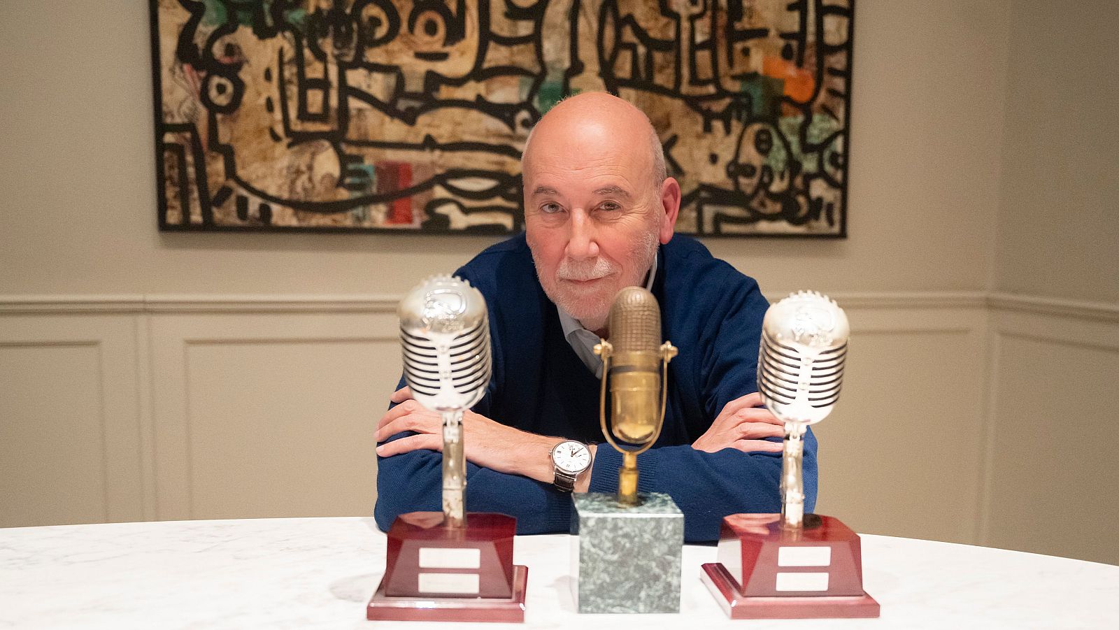 Màrius Carol amb 3 premis en forma de micròfon, assegut amb els braços plegats i una mirada directa a càmera.