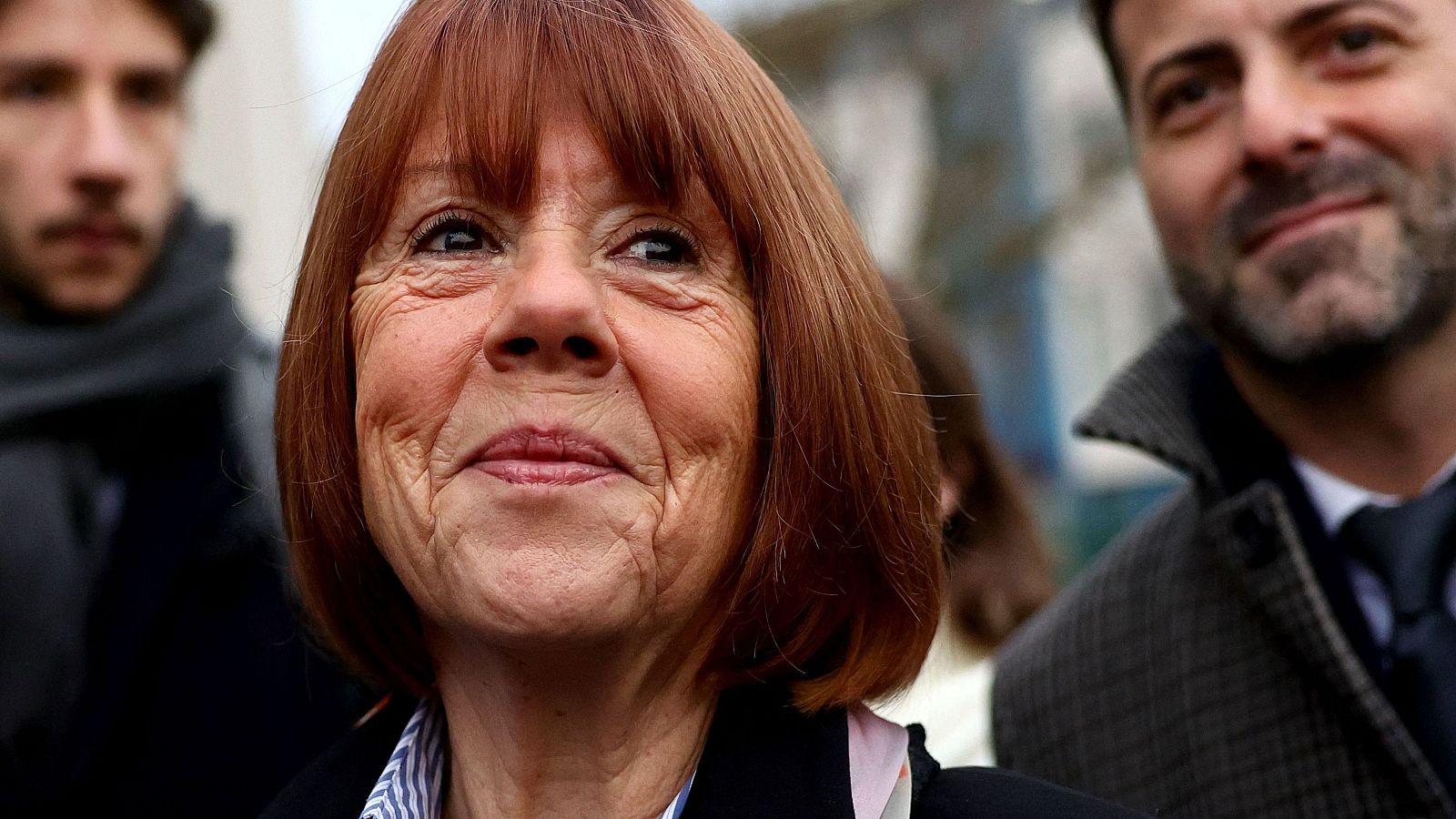 Gisèle Pelicot, rostro arrugado y pelo corto rojizo, mira hacia arriba con expresión serena y satisfecha.  Fondo desenfocado con personas.