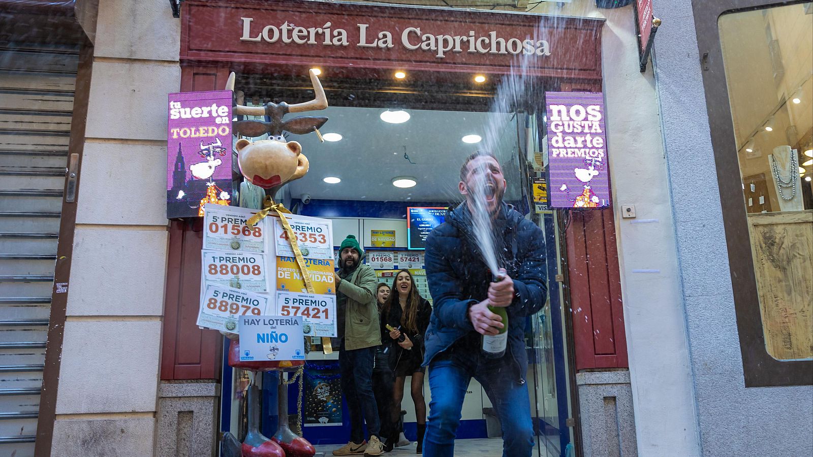Un hombre celebra efusivamente un premio de lotería descorchando champán en la administración "La Caprichosa", donde se ven carteles con números premiados (ej: 88008, 01568) y otros anuncios.