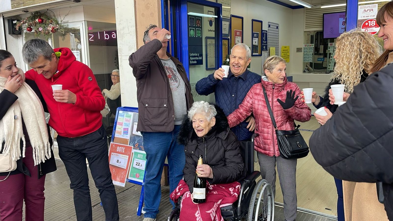 Celebración festiva tras un premio de lotería.  Brindis, alegría y una mujer en silla de ruedas con una botella de cava. Cartel con números 8 y 22.