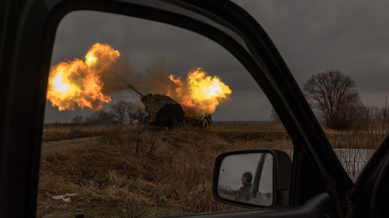 Disparo de tanque en paisaje rural; dos llamaradas anaranjadas; vista desde vehículo con reflejo en retrovisor.