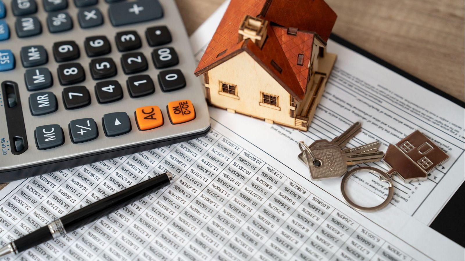 Objetos que simbolizan la compra de una vivienda (casa, llaves, calculadora) sobre un documento con datos numéricos, ilustrando una noticia sobre hipotecas más baratas en 2025.