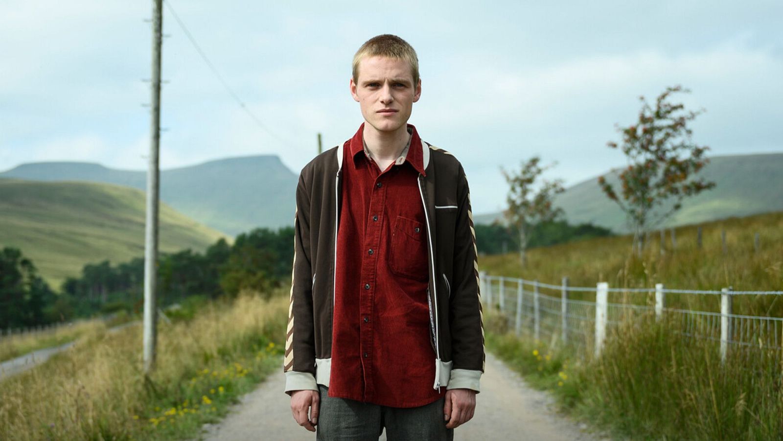 Joven con chaqueta marrón y camisa roja en camino rural, paisaje montañoso y cielo nublado al fondo.