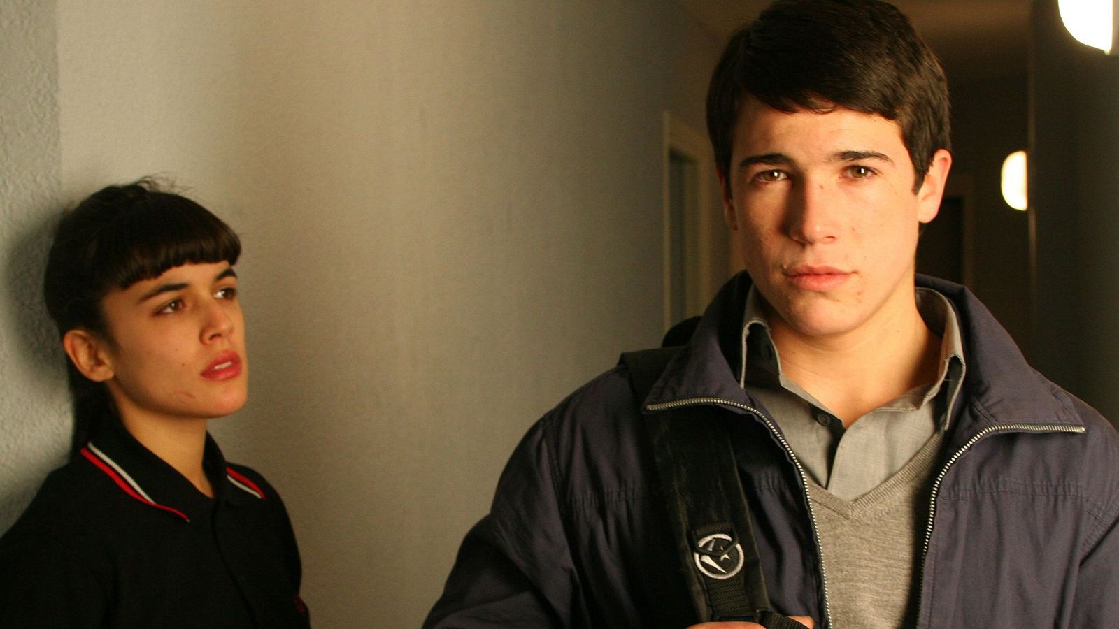 Jóvenes en pasillo, él con chaqueta azul y mochila, ella con polo negro. Iluminación en rostros. Posible escena de película.