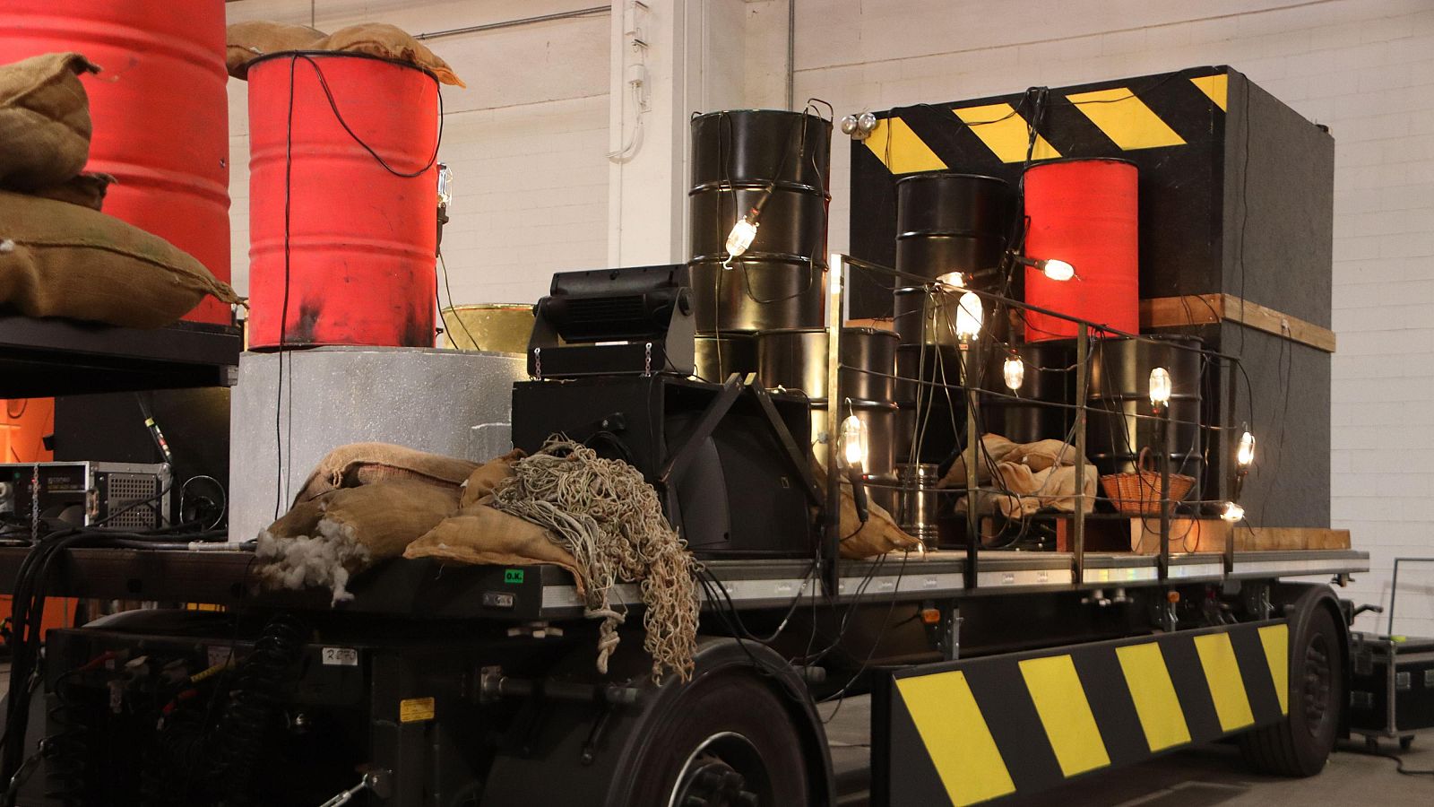 Remolque con barriles, cajas y sacos; franjas amarillas y negras de advertencia; posible instalación eléctrica temporal.