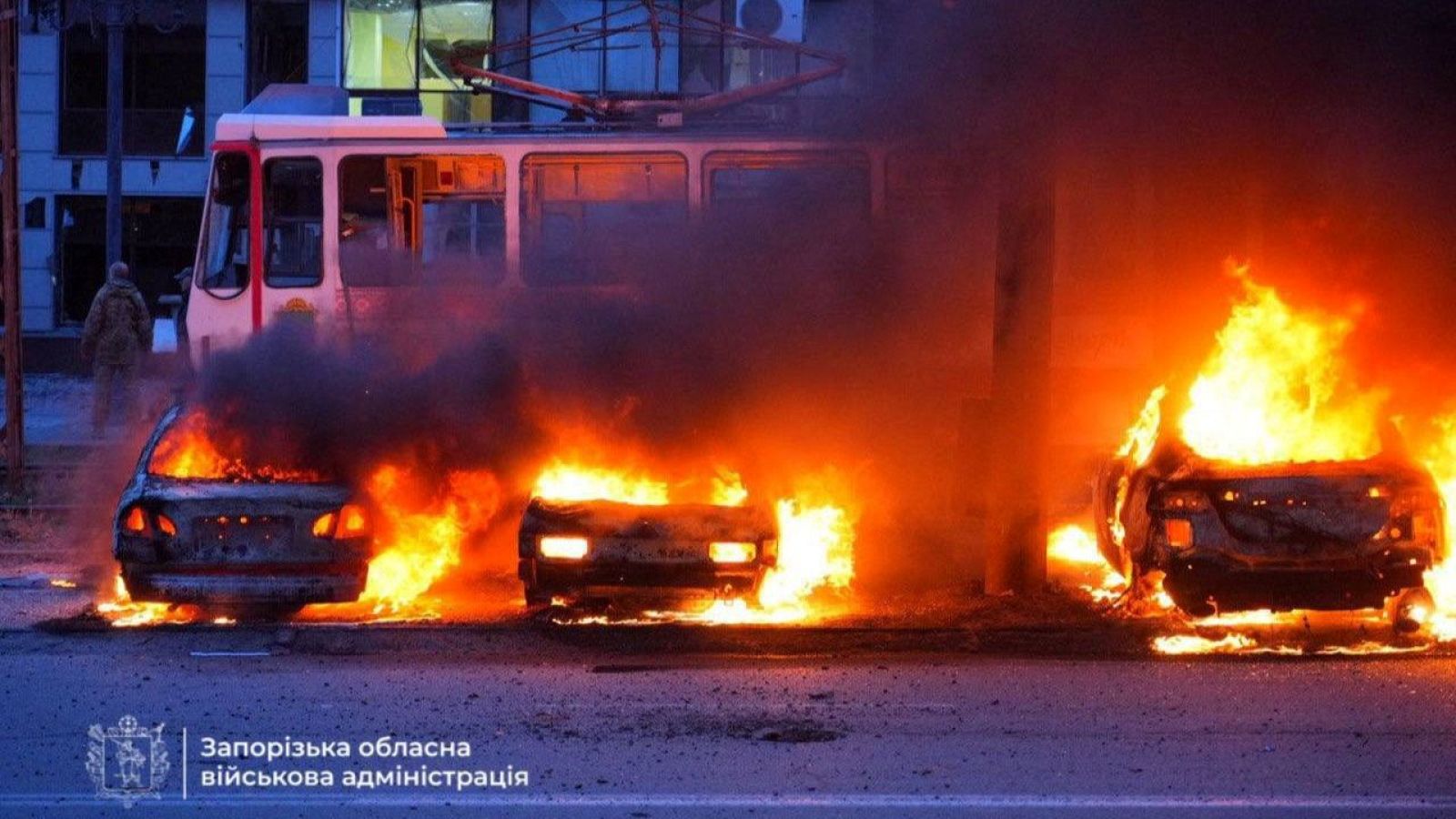 Coches en llamas tras un ataque; un tranvía cercano parece intacto; una persona en uniforme o ropa similar está presente.