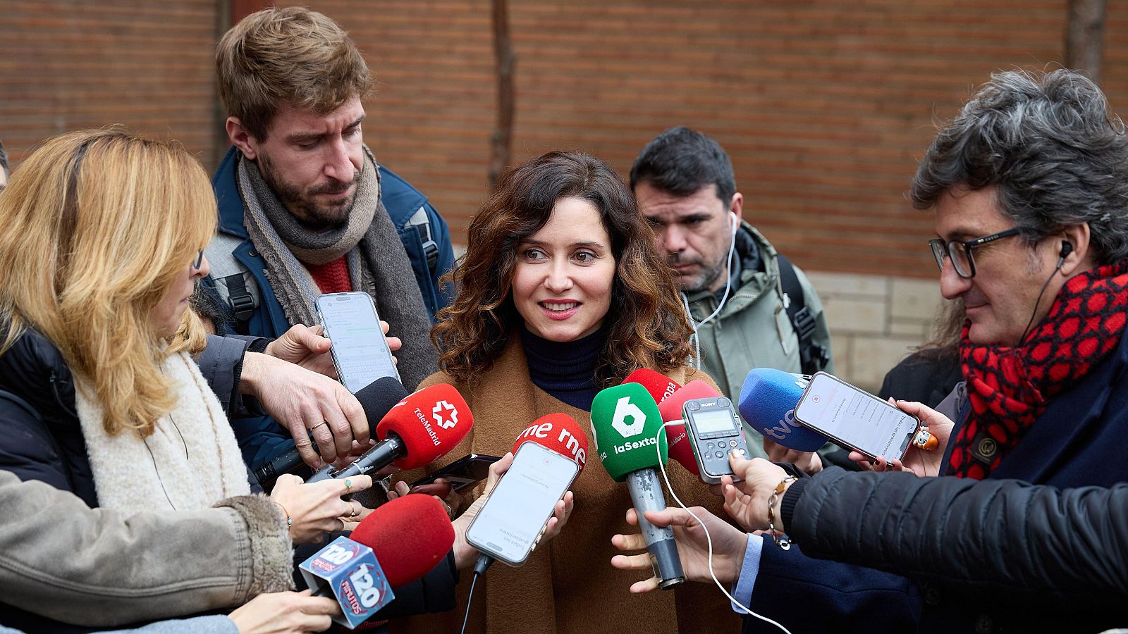 La presidenta de la Comunidad de Madrid atiende a los medios, rodeada de periodistas con micrófonos y grabadoras en un acto público exterior.