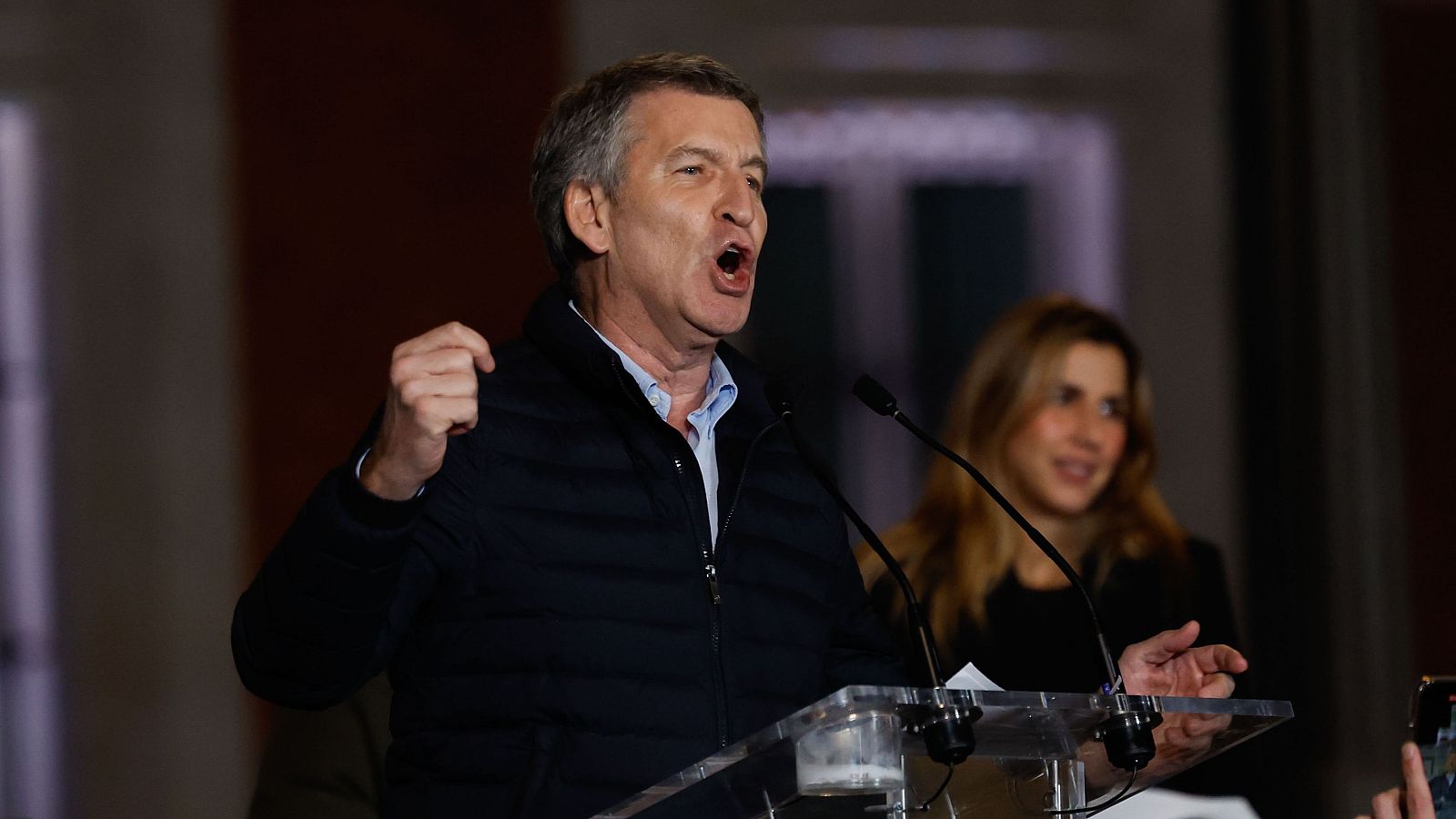 Imagen de Alberto Núñez Feijóo dando un discurso con gesto enérgico; lleva chaqueta oscura y camisa clara; una mujer con cabello rubio está detrás.