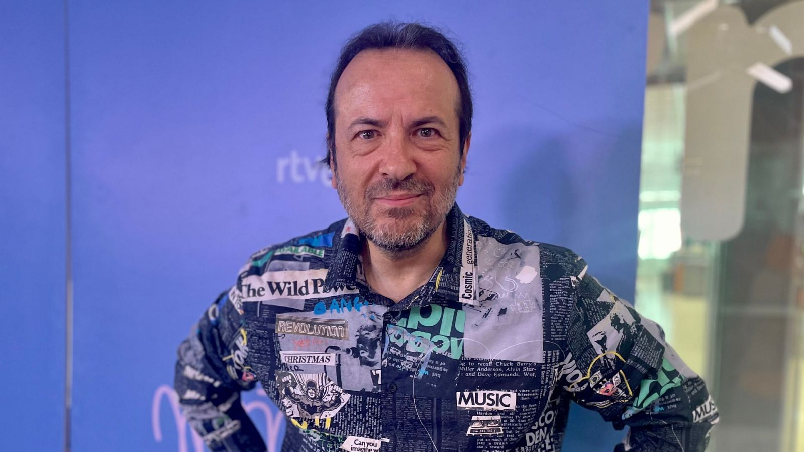Hombre de mediana edad con camisa estampada con recortes de periódico y palabras como "Revolution" y "Christmas". Fondo azul liso y reflejo en cristal.