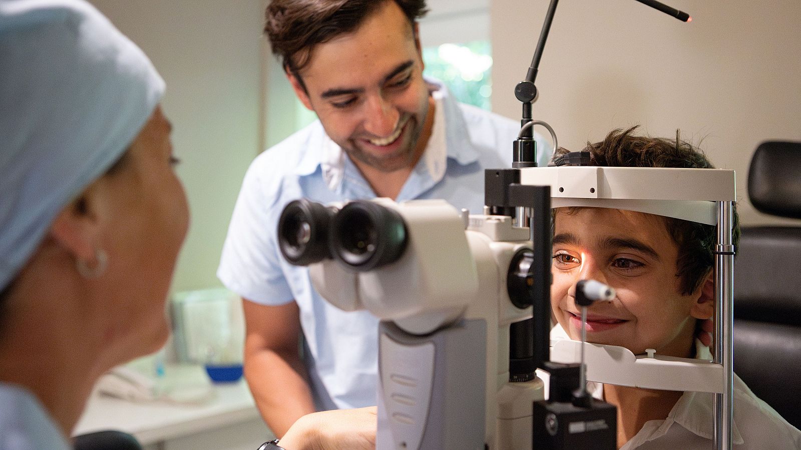 Revisión oftalmológica infantil: niño sonriente durante examen de la vista, supervisado por oftalmólogo y asistente.