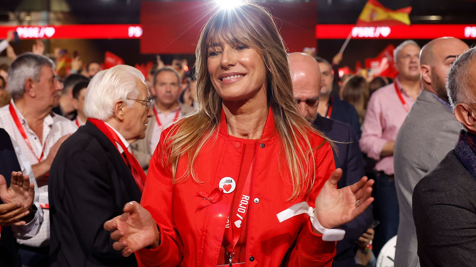 Begoña Gómez, con chaqueta roja, sonríe y aplaude en un ambiente festivo.  Se aprecian banderas y pancartas al fondo.
