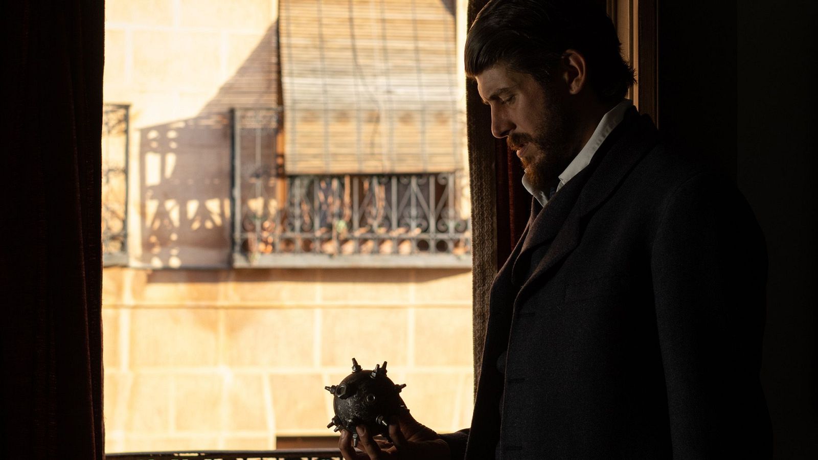 Hombre con traje oscuro y camisa blanca, sosteniendo una bomba esférica cerca de una ventana con reja.  Expresión seria. Imagen en blanco y negro.