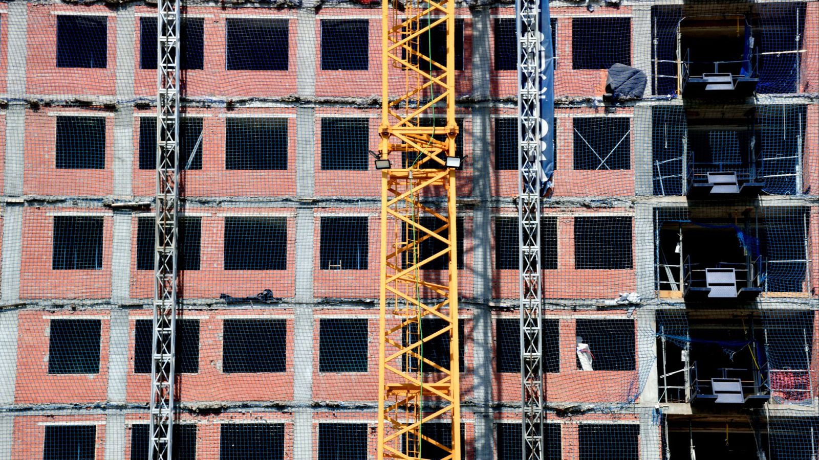 Edificio en construcción con estructura de ladrillo visto, ventanas protegidas con malla y grúa amarilla presente. Andamios metálicos visibles.