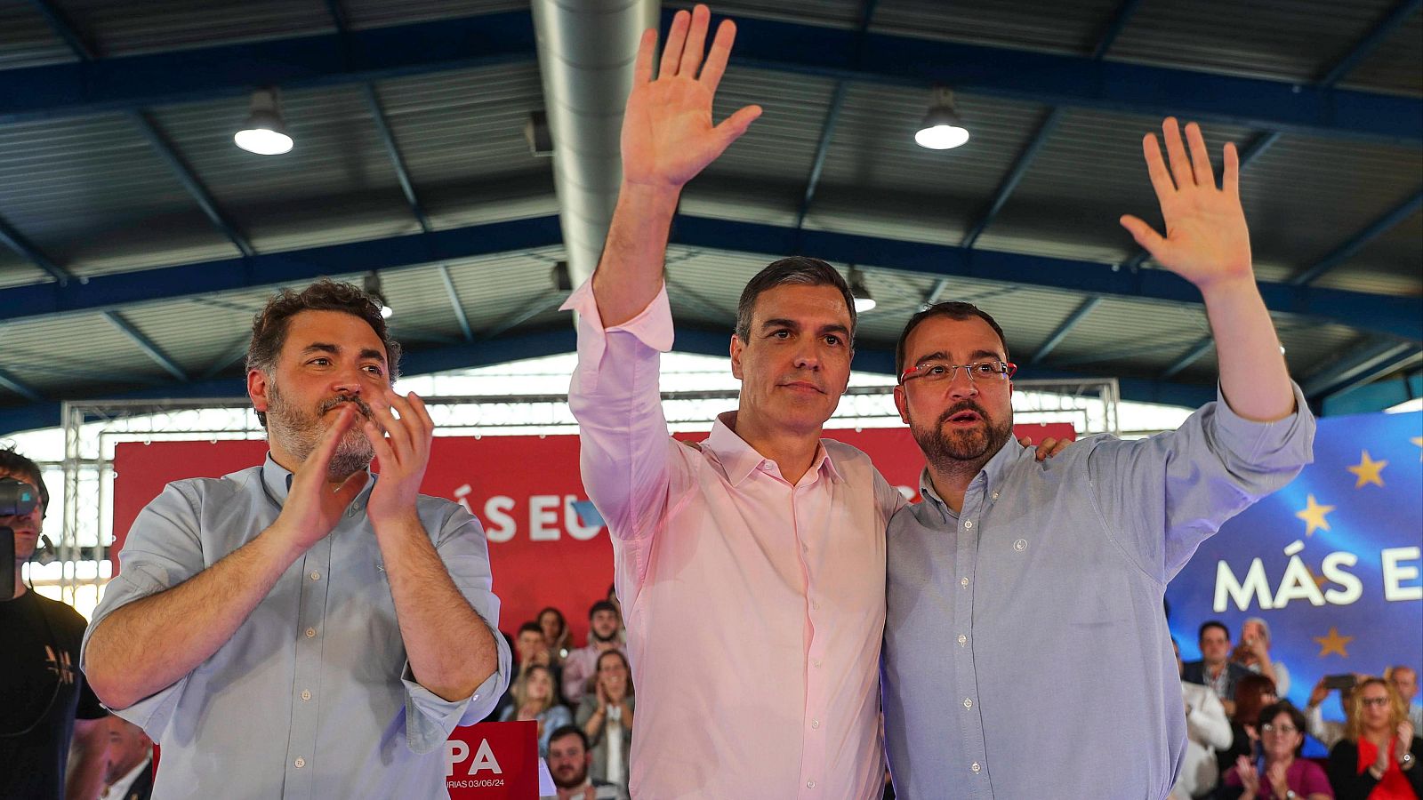 Saludo de Pedro Sánchez y Adrián Barbón a un público que aplaude en un evento de 2024.  Ambos llevan camisas de colores claros; se ve un cartel con un mensaje político.