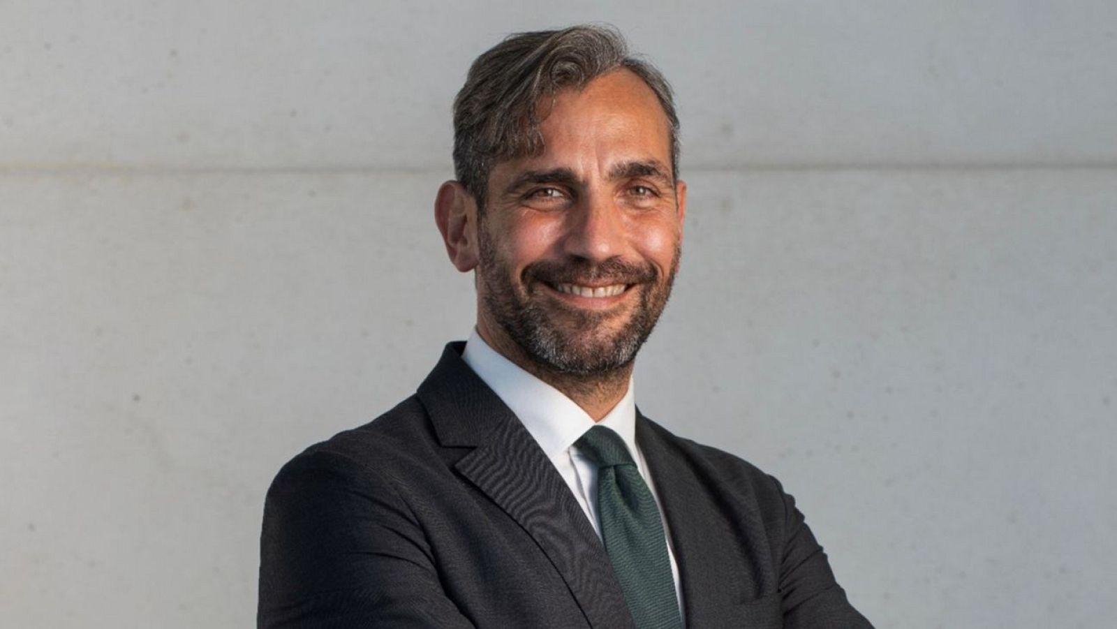 Retrato de un hombre de mediana edad con traje oscuro, camisa blanca y corbata verde, sonriendo con los brazos cruzados sobre un fondo gris claro.