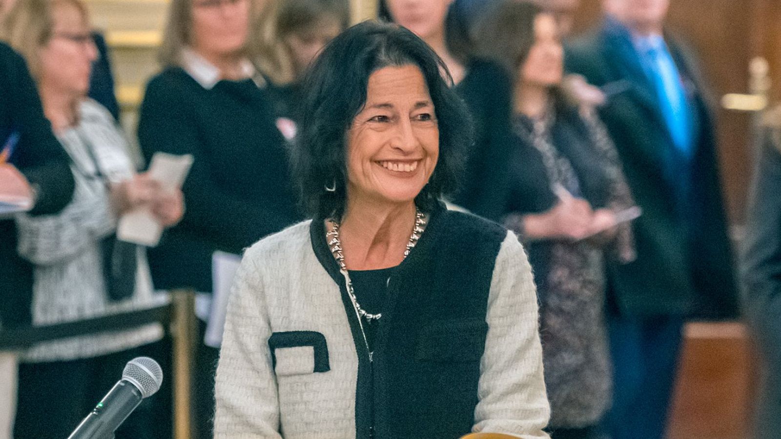 Mercedes de Pablos, en lo que parece una rueda de prensa, sonríe y mira a la derecha. Lleva chaqueta bicolor y collar plateado; un micrófono está en primer plano.