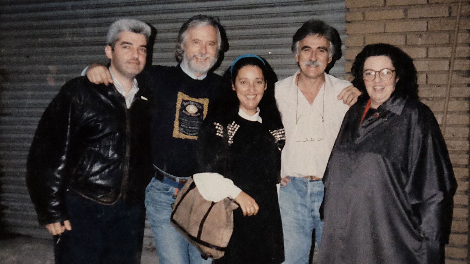 Grupo de cinco personas (cuatro hombres y una mujer) posando informalmente, con vestimenta casual y expresiones amigables, frente a una puerta enrollable metálica.