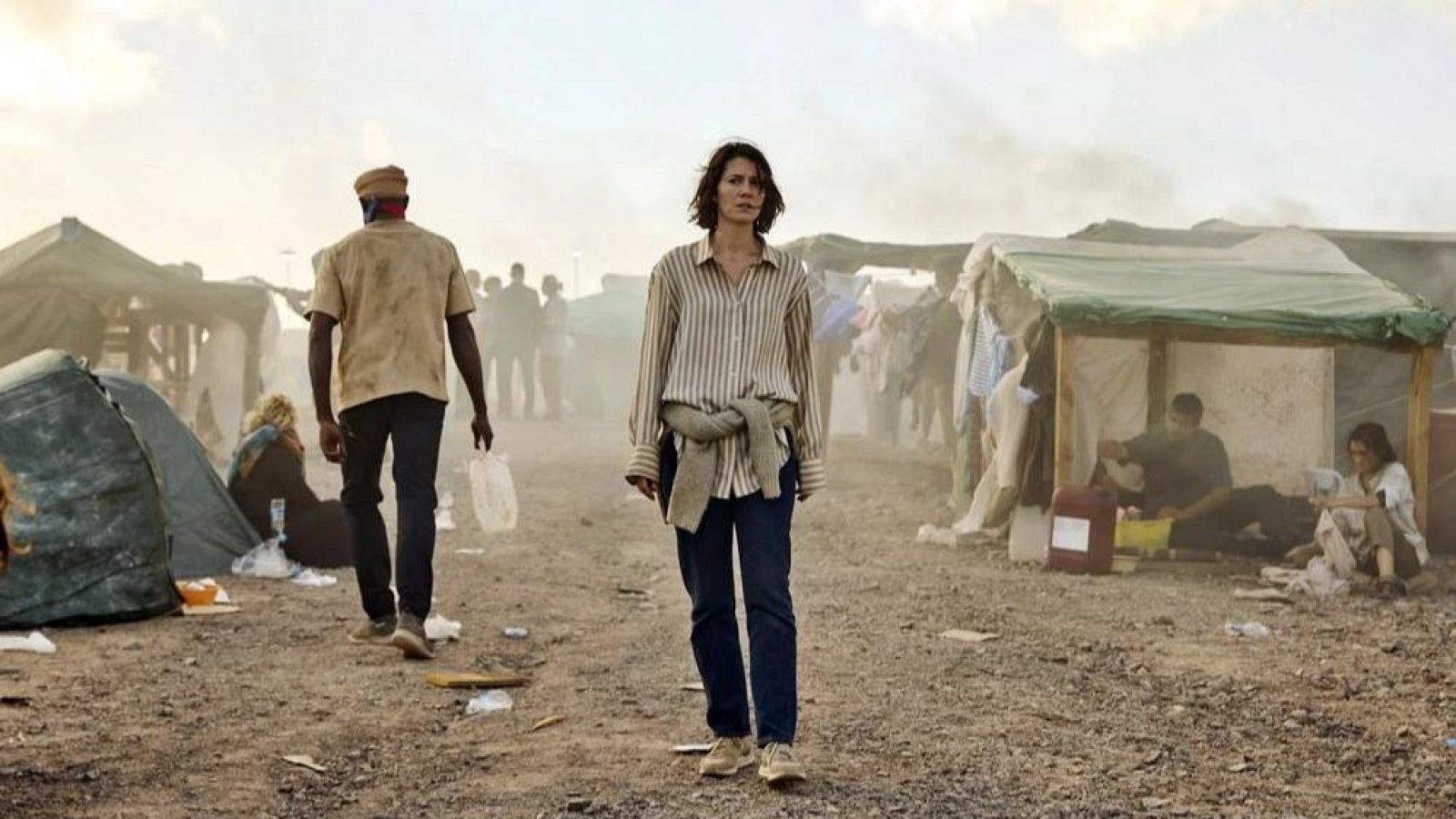 Mujer caminando hacia la cámara en un campamento pobre; un hombre se aleja.  Ropa sencilla, ambiente desolado.  Contrastes sociales.