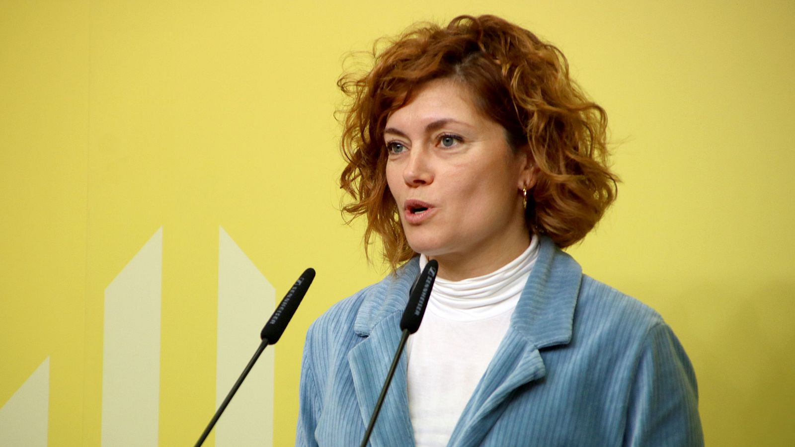 Portavoz de ERC ofreciendo una rueda de prensa. Lleva chaqueta azul y jersey blanco, hablando a dos micrófonos sobre un fondo amarillo con líneas verticales.