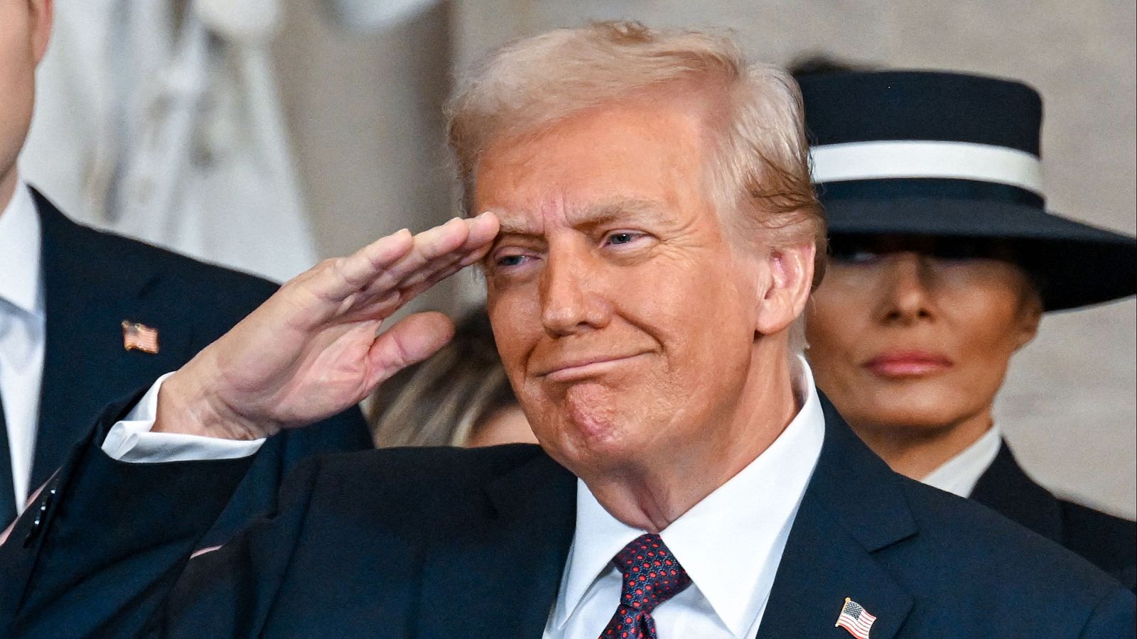 Primer plano de Donald Trump saludando militarmente en su investidura. Viste traje oscuro, camisa blanca y corbata roja con lunares blancos. Melania Trump aparece al fondo.