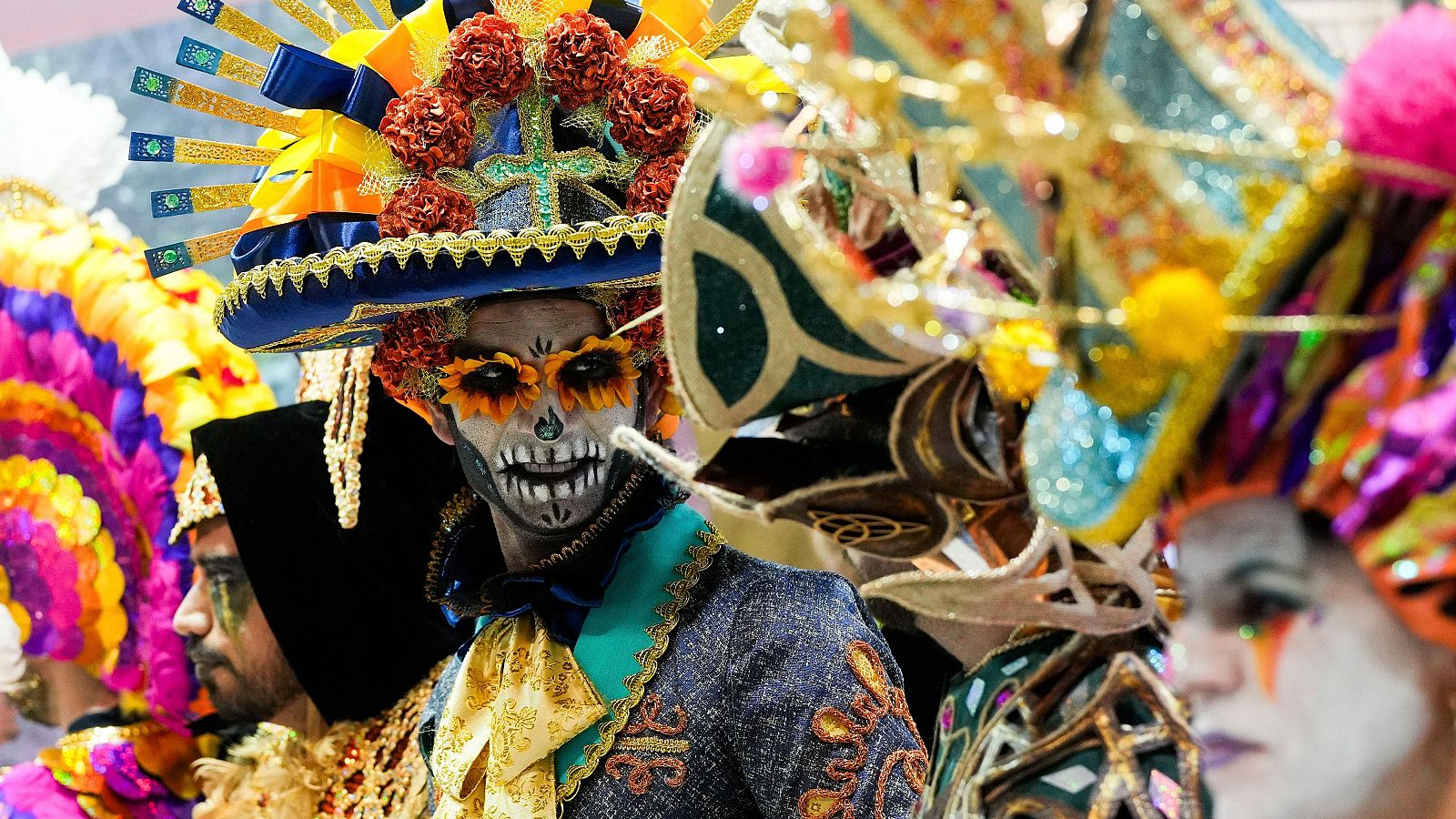 Diversidad de trajes y tocados coloridos en un evento turístico. Una persona lleva maquillaje de calavera y sombrero decorado.  Amplia representación cultural.