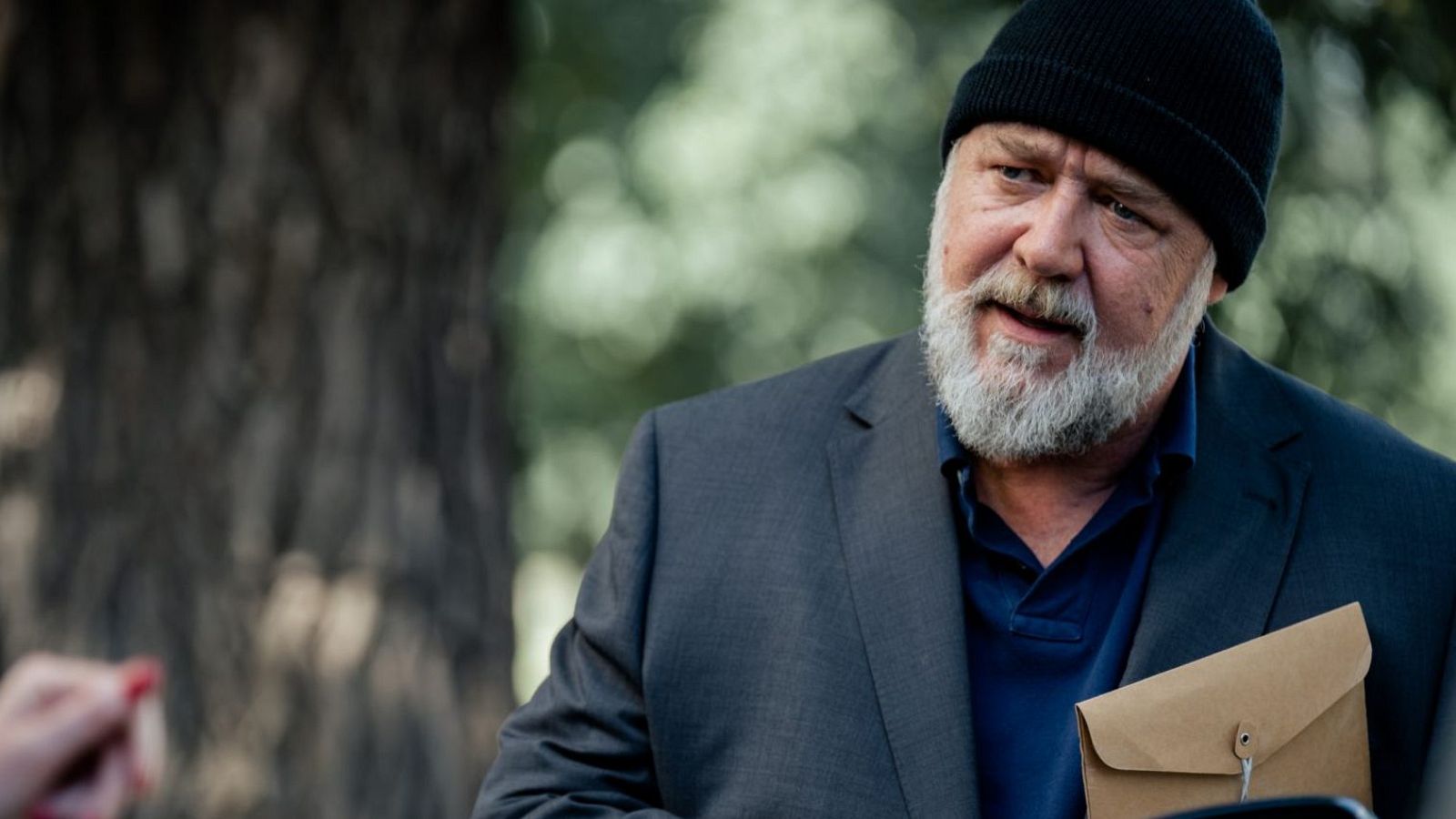 Juan Echanove, con barba blanca y gorro, sostiene un sobre marrón en un entorno exterior.  Americana gris y camisa azul marino.