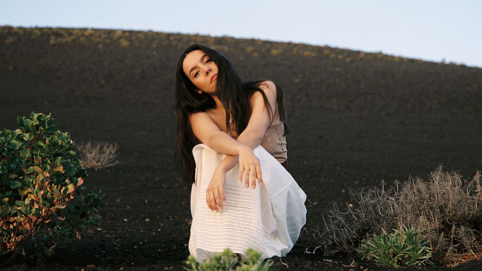 Cantante con top beige y falda larga blanca sentada en terreno volcánico, mirando a cámara con expresión neutra.  Atardecer.