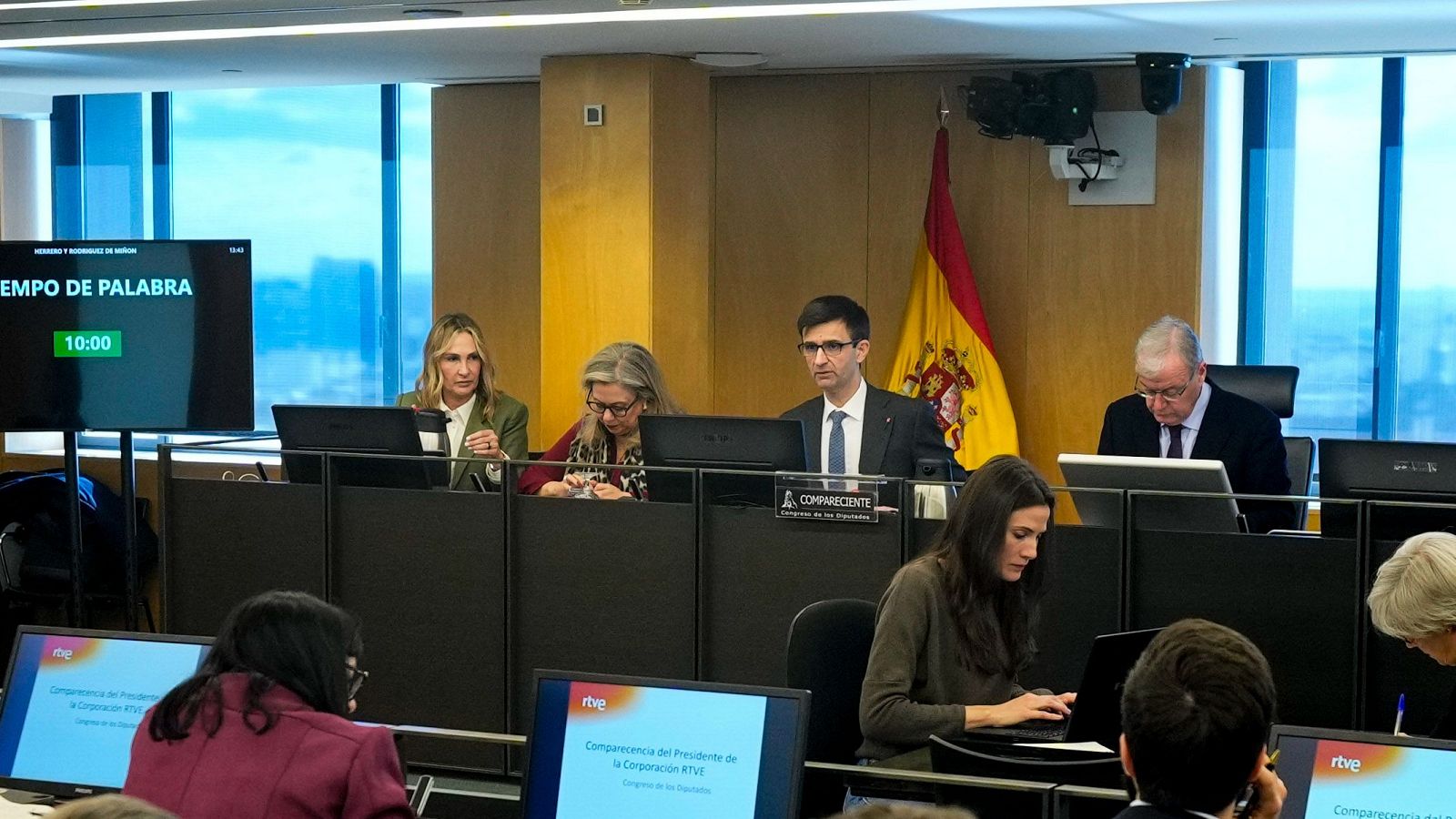 Comparecencia de un hombre con traje y gafas ante una comisión, en una sala con ordenadores, micrófonos y una pantalla mostrando 'TIEMPO DE PALABRA 10:00'.