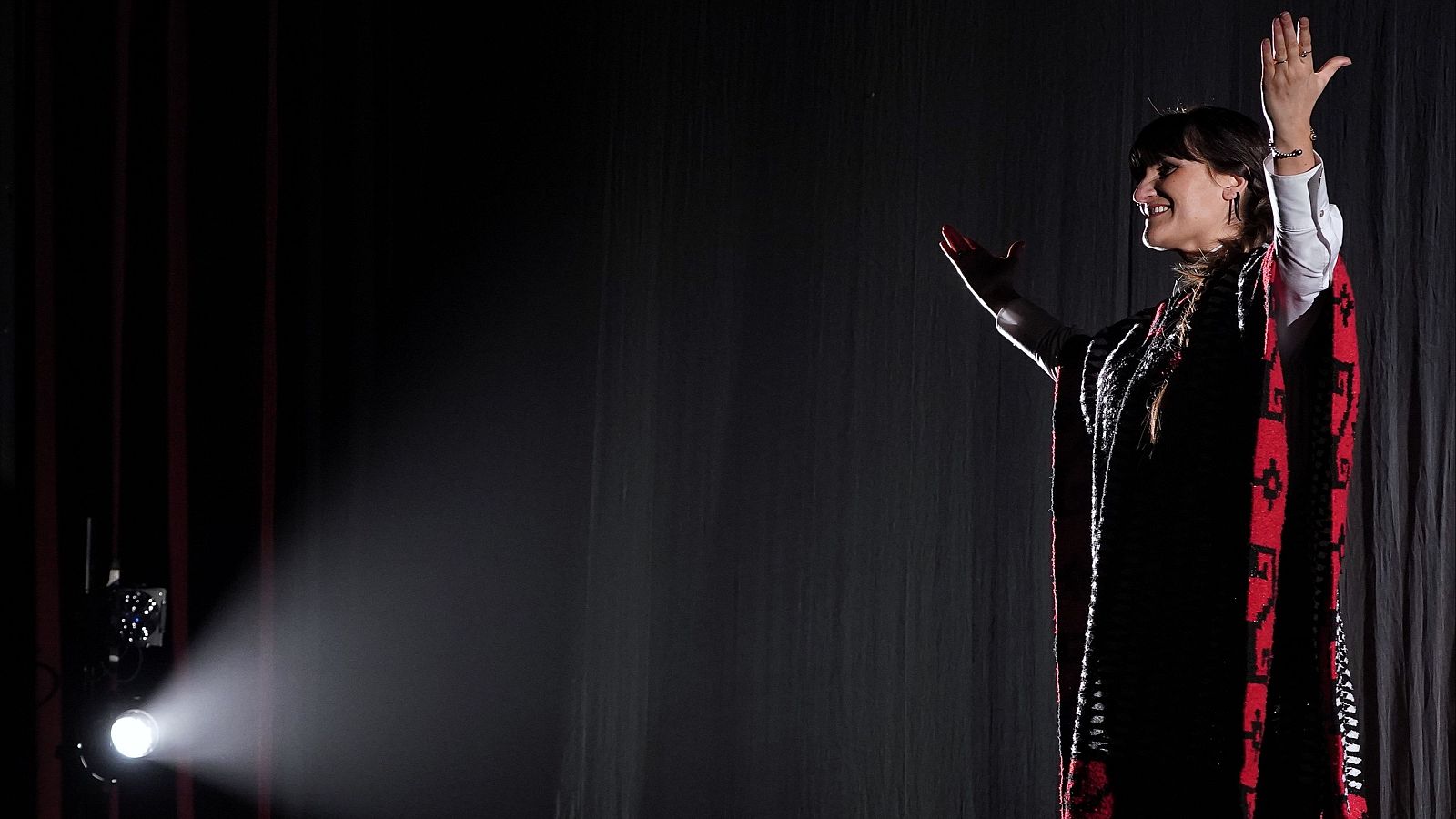 Actriz en escena teatral, iluminada, con poncho oscuro y detalles rojos, brazos extendidos, expresión alegre. Posiblemente de la obra "Chavela".