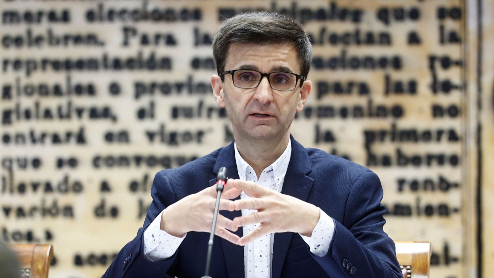 Hombre con traje oscuro y gafas habla ante un micrófono en una sesión formal. Gestos con las manos. Fondo con texto ilegible.