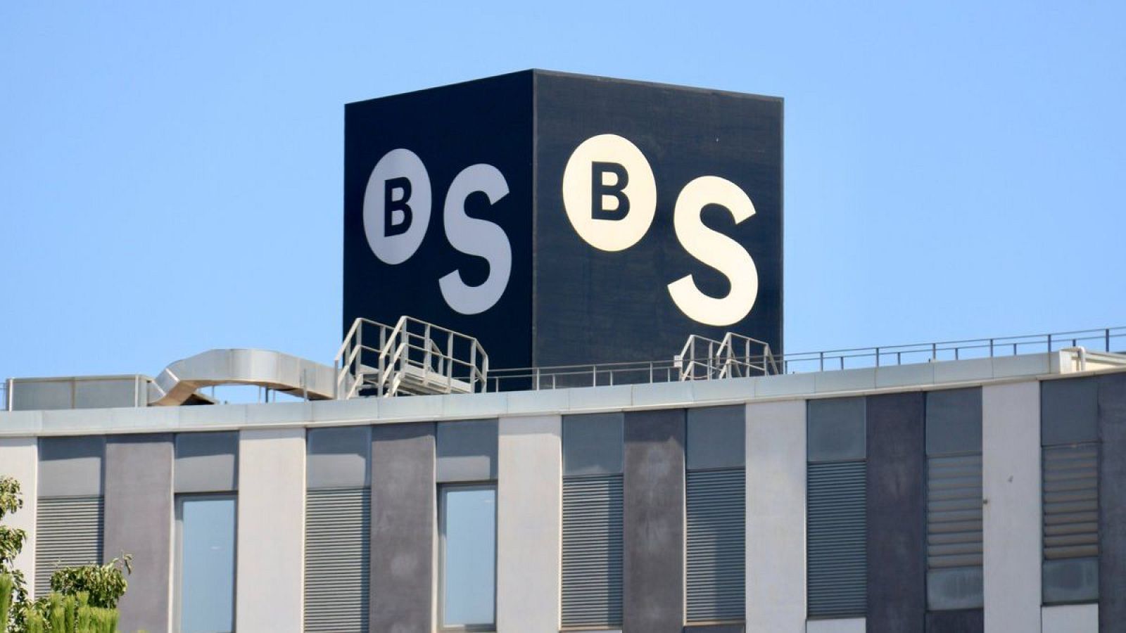 Logotipo "BS" blanco sobre fondo negro en la azotea de un edificio moderno con fachada gris y blanca. Se observan ventanas y estructuras metálicas.