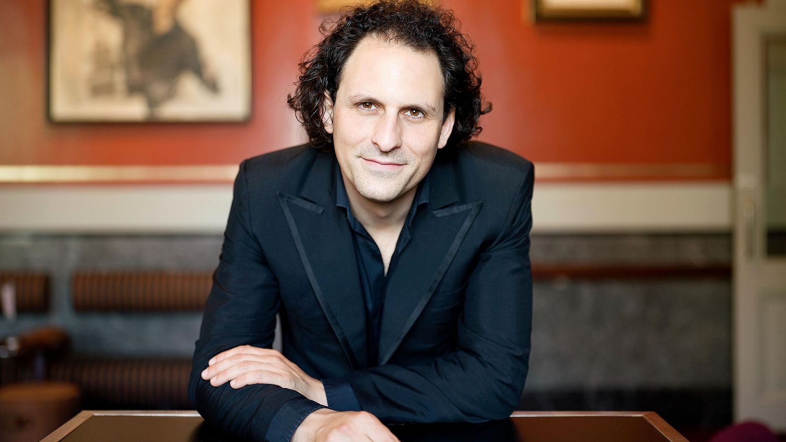 Retrato de Jone Martínez: director de orquesta sentado, americana negra, pelo rizado oscuro, fondo con paredes rojizas y cuadros.