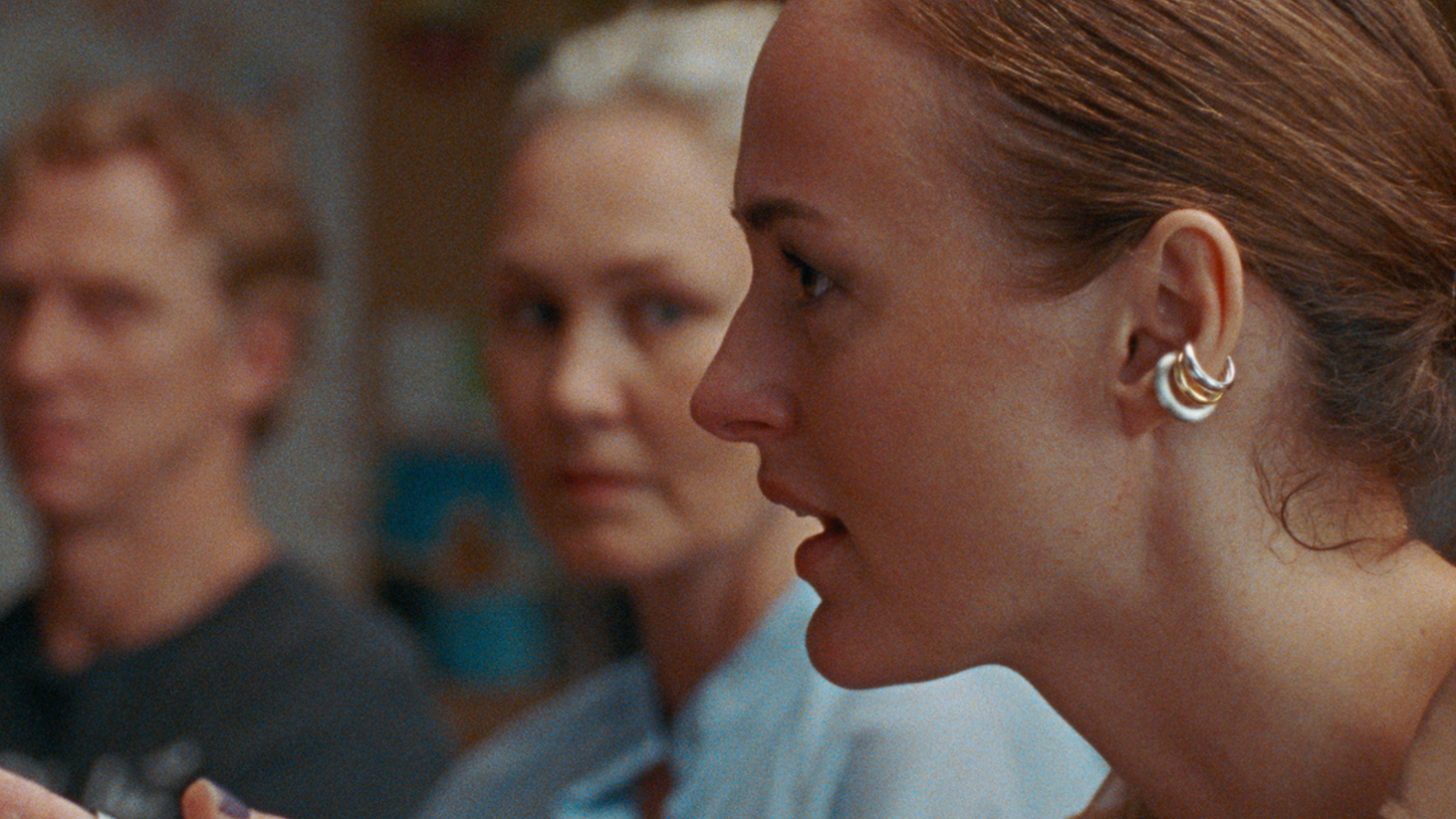 Primer plano del perfil de una joven con pendientes; al fondo, dos personas más, una mujer y un hombre, ligeramente desenfocadas. Escena interior.