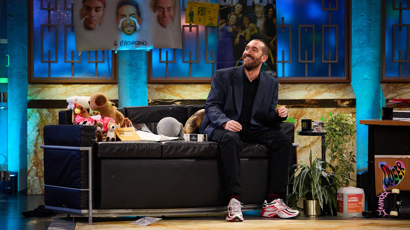 Humorista sentado en un sofá, sonriendo, con chaqueta oscura y pantalones negros.  En el set hay peluches, una taza y una planta.