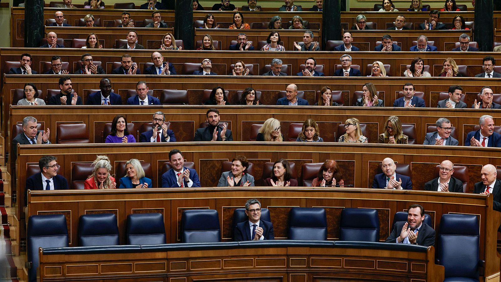 Hemiciclo del Congreso de los Diputados durante una sesión; numerosos diputados aplauden en sus escaños de madera con tapicería azul oscuro.
