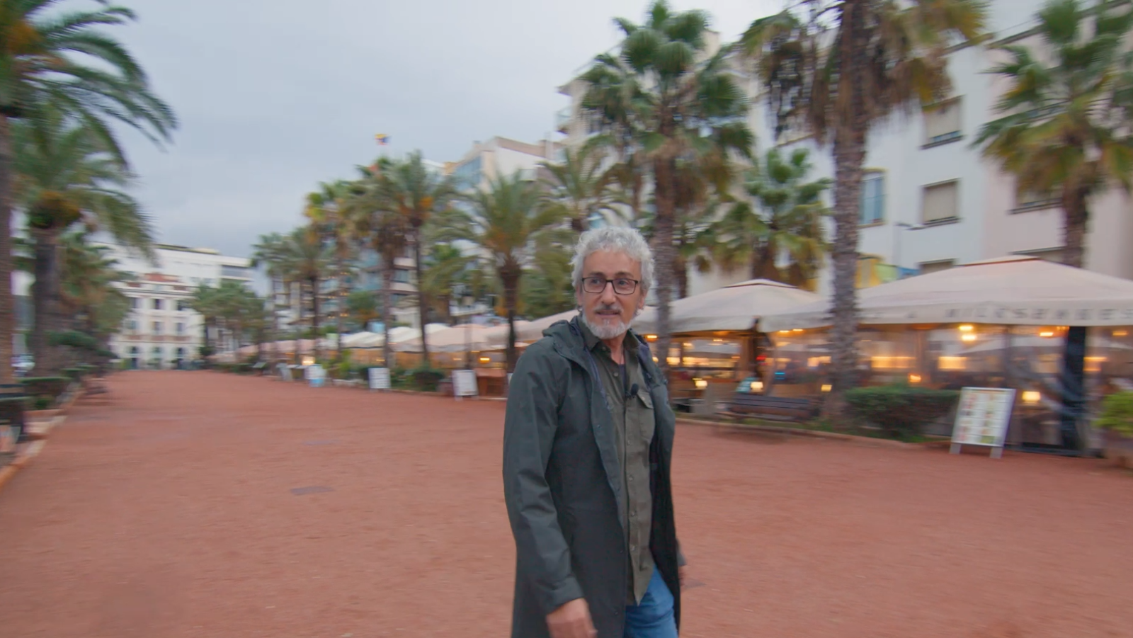 Presentador con chaqueta oscura y camisa verde oliva caminando por calle peatonal con palmeras y locales de hostelería. Iluminación tenue, pavimento rojizo.